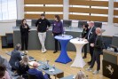 Man sieht eine Gruppe Menschen, die sich miteinander austauschen. Vor ihnen stehen ein blauer und ein weißer Stehtisch. In der unteren Bildhälfte sieht man das Publikum von hinten.